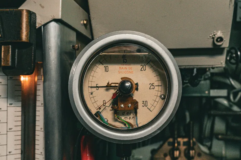 Analog mätare monterad bland rör och kablar i en industrimiljö.